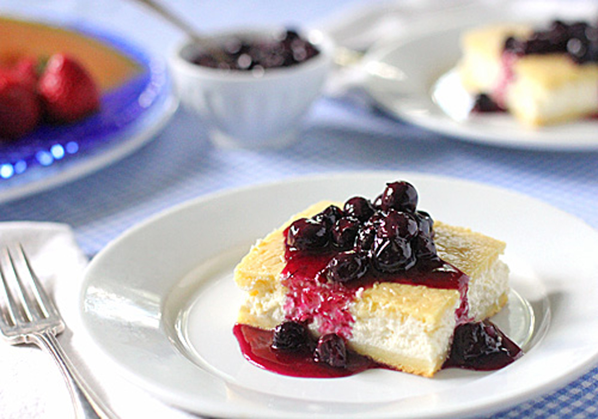 A slice of cheesecake topped with blueberry sauce sits on a white plate, with a fork beside it. In the background, a bowl of blueberry sauce and another dessert plate are visible.