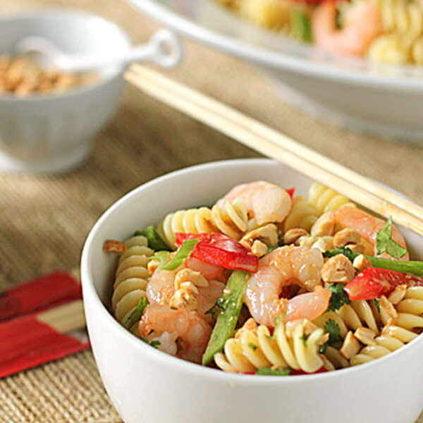A bowl of rotini pasta salad with shrimp, red bell peppers, green beans, chopped nuts, and herbs. Chopsticks rest on the bowl, and additional servings appear in the background.