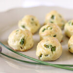 A plate with several golden-brown, round pastry puffs with green herbs visible inside, displayed next to two purple chive flowers and green stems on a light-colored plate.