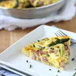 A slice of frittata with ham, asparagus, and chives sits on a square white plate with a fork. A purple chive flower and extra chives garnish the plate; the rest of the frittata is visible in the background.