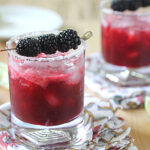 Two glasses of a dark red cocktail with ice, garnished with blackberries on a skewer and a sugared rim, sit on patterned napkins. Lime wedges and extra blackberries are scattered nearby.