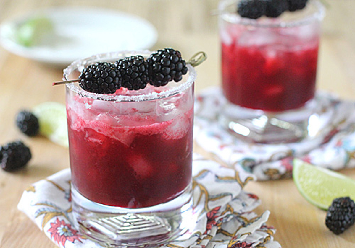 Two glasses of a dark red cocktail with ice, garnished with blackberries on a skewer and a sugared rim, sit on patterned napkins. Lime wedges and extra blackberries are scattered nearby.