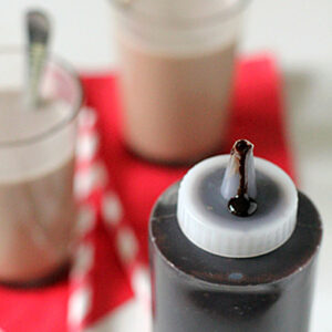 A bottle of chocolate syrup with a drop on the tip is in focus, positioned in front of two glasses of chocolate milk on a red napkin, with white and red striped straws beside them.