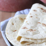 A close-up of soft, folded flour tortillas stacked on a plate lined with a blue cloth, with more tortillas blurred in the background.