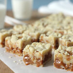 Close-up of caramel crumb bars cut into squares on parchment paper, with two glasses of milk in the background on a wooden surface.