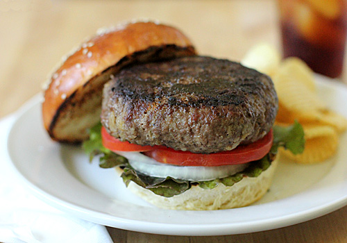 A hamburger with a thick beef patty, lettuce, tomato, and onion sits on an open bun on a white plate, with potato chips on the side.