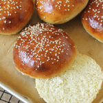Golden brown sesame seed hamburger buns on a baking tray, with one bun sliced open to show its soft, fluffy interior.