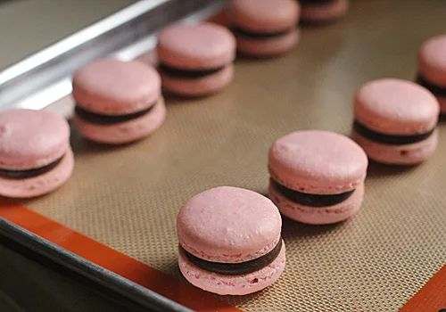 Raspberry Chocolate French Macarons with pink shells and chocolate filling are arranged on a baking tray lined with a silicone baking mat.