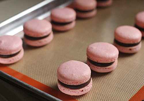 Raspberry Chocolate French Macarons with pink shells and chocolate filling are arranged on a baking tray lined with a silicone baking mat.