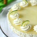 A creamy, round key lime cheesecake topped with whipped cream swirls and lime slices, with coconut flakes around the sides, displayed on a glass cake stand.