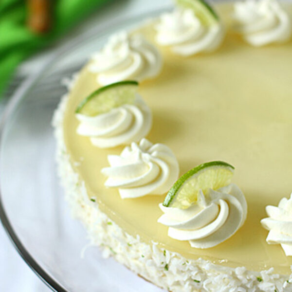 A creamy, round key lime cheesecake topped with whipped cream swirls and lime slices, with coconut flakes around the sides, displayed on a glass cake stand.