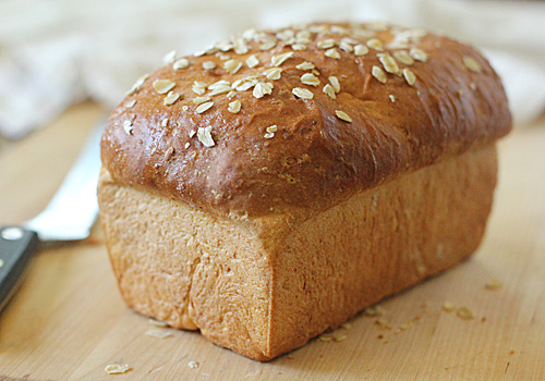 A loaf of golden-brown bread topped with rolled oats sits on a wooden surface, with a knife partly visible in the background.