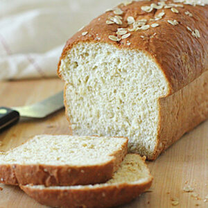 A loaf of oat bread sits on a wooden surface with two slices cut in front. A bread knife rests nearby, and a white cloth is in the background. Oats are sprinkled on top of the bread.