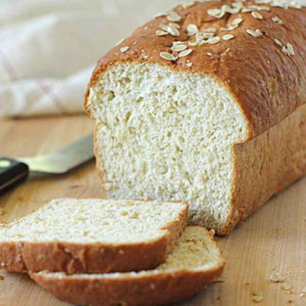 A loaf of oat bread sits on a wooden surface with two slices cut in front. A bread knife rests nearby, and a white cloth is in the background. Oats are sprinkled on top of the bread.
