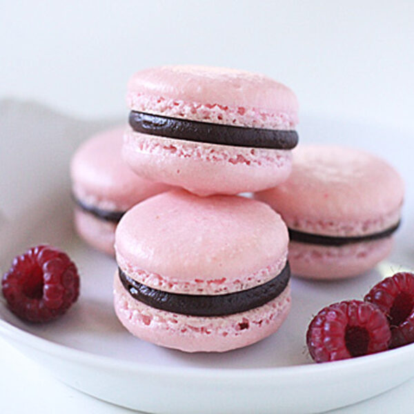 Four pink macarons with dark chocolate filling are stacked on a white plate, accompanied by fresh raspberries. The background is plain white, giving a clean and elegant look to the arrangement.