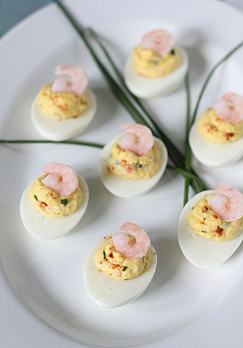 Deviled eggs arranged on a white plate, each topped with a small shrimp and sprinkled with seasoning. Green chives are placed decoratively in the center.