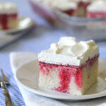 A square slice of white poke cake with red strawberry filling and whipped cream topping sits on a white plate, with a fork beside it, on a blue checkered tablecloth. More cake is visible in the background.