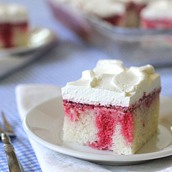 A square slice of white poke cake with red strawberry filling and whipped cream topping sits on a white plate, with a fork beside it, on a blue checkered tablecloth. More cake is visible in the background.