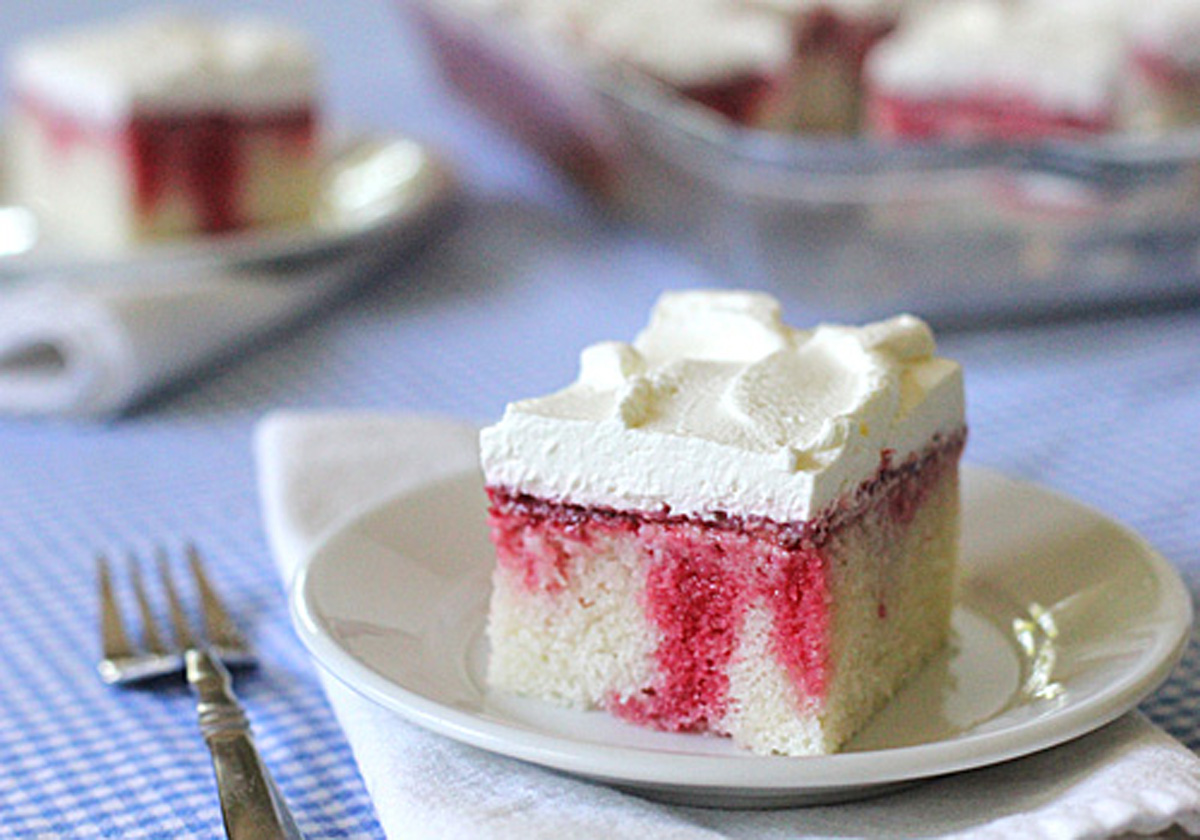 A square slice of white poke cake with red strawberry filling and whipped cream topping sits on a white plate, with a fork beside it, on a blue checkered tablecloth. More cake is visible in the background.