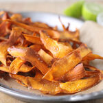 A close-up of crispy, salted sweet potato chips on brown parchment paper in a metal tray, with lime wedges and a small bowl in the blurred background.