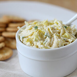 A white bowl filled with creamy dip, garnished with herbs, sits next to a plate of round crackers. A white spoon rests in the dip. The bowl and crackers are on a white plate with a neutral cloth nearby.