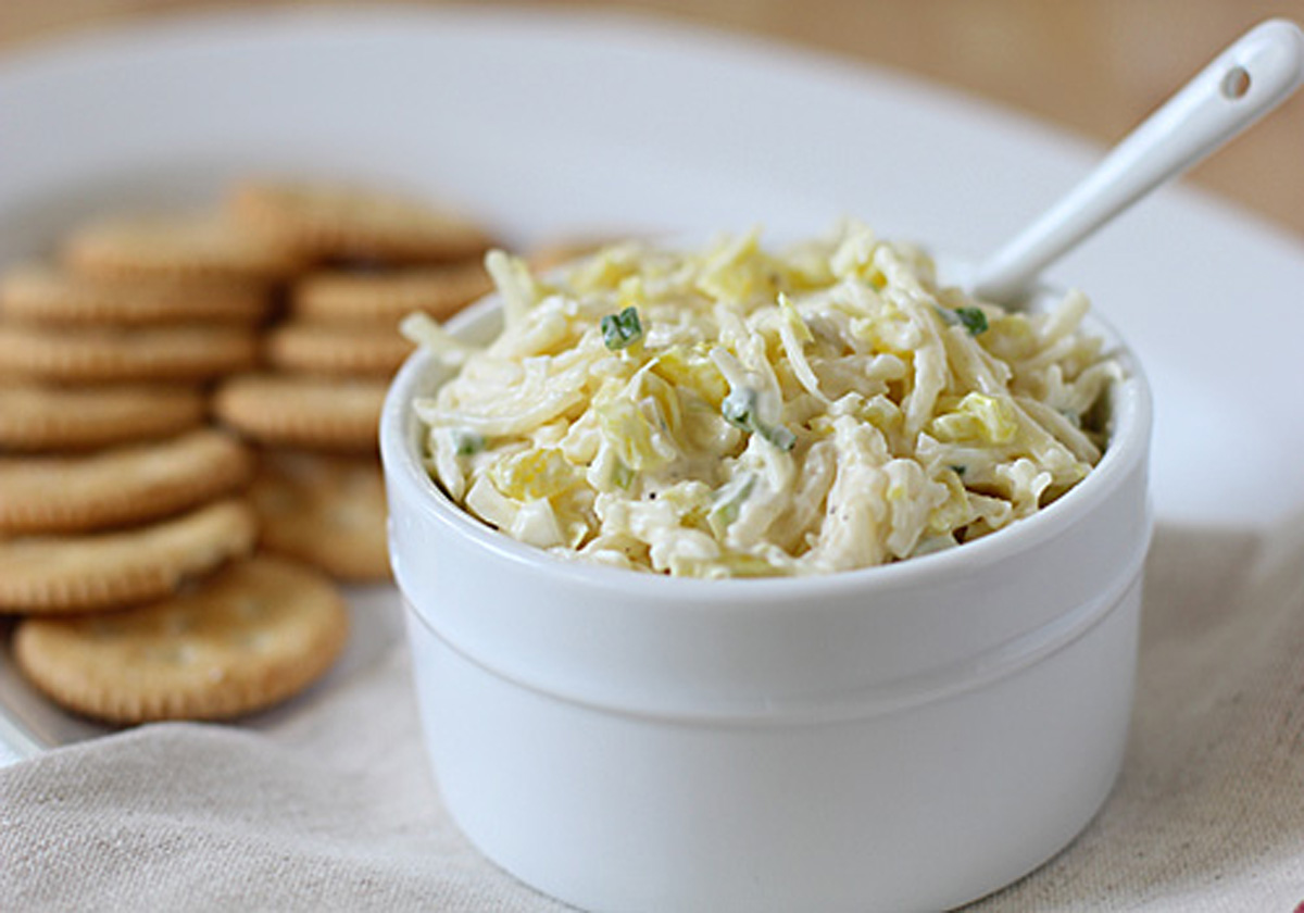 A white bowl filled with creamy dip, garnished with herbs, sits next to a plate of round crackers. A white spoon rests in the dip. The bowl and crackers are on a white plate with a neutral cloth nearby.