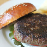 A close-up of a hamburger on a white plate, with a sesame seed bun, lettuce, tomato, onion, and a thick beef patty. The top bun is slightly tilted to reveal the layers inside the burger.