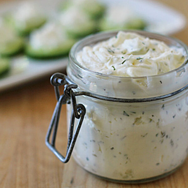 A glass jar filled with herbed cream cheese sits on a wooden surface, with cucumber slices topped with the same spread visible on a plate in the background.