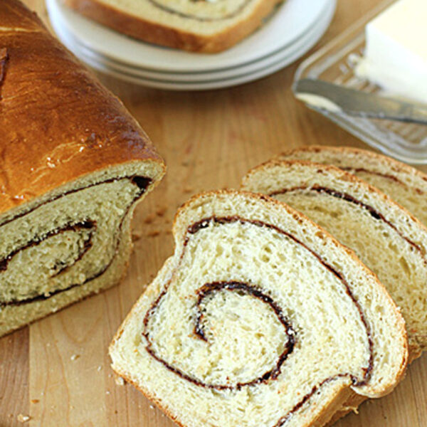 Sliced cinnamon swirl bread on a wooden surface, with a loaf, a butter dish, a knife, and two plates of buttered bread visible in the background.