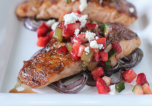 A piece of cooked salmon topped with a colorful salsa of diced strawberries, cucumber, and crumbled cheese, served on a bed of roasted red onion slices.
