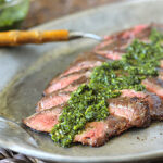 Sliced medium-rare steak arranged on a gray plate, topped with a green chimichurri sauce, with a serving utensil and a bowl of extra sauce in the background.