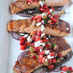 Grilled salmon fillets are served on a bed of sliced red onions, topped with a colorful mix of diced cucumbers, tomatoes, herbs, and crumbled feta cheese on a white platter.