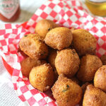 A basket lined with red and white checkered paper filled with golden-brown hush puppies, with a bottle of hot sauce and a bottle of beer in the background.