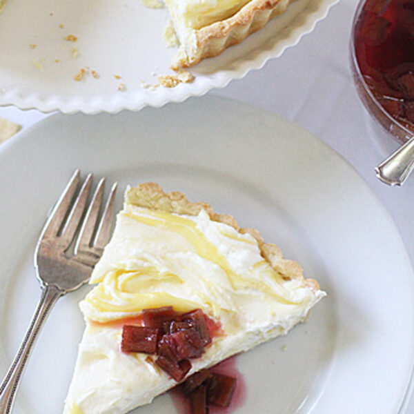 A slice of creamy lemon tart topped with rhubarb compote is served on a white plate with a fork. The rest of the tart and a bowl of compote are visible nearby.