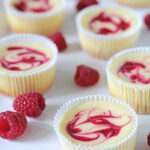 Mini cheesecakes in white cupcake liners, each topped with a red raspberry swirl, are arranged on a white surface with fresh raspberries scattered around them.