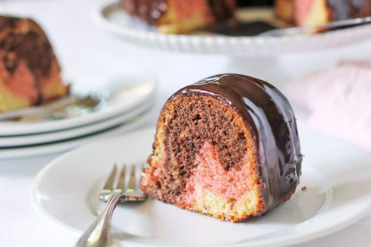 A slice of neapolitan bundt cake with chocolate glaze is on a white plate with a fork. The cake features irresistible chocolate, pink, and yellow swirls reminiscent of classic Neapolitan flavors. Additional cake slices are in the background.