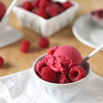A white bowl filled with scoops of raspberry sorbet, garnished with fresh raspberries and a white spoon. In the background, there are more raspberries and a white container on a wooden table.