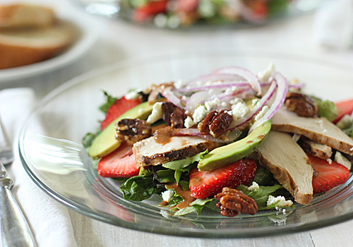 A glass plate of salad with sliced chicken, strawberries, avocado, red onion, candied pecans, and crumbled cheese on greens. Slices of bread are blurred in the background.