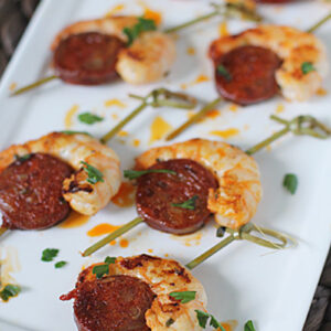 Skewers with grilled shrimp and slices of sausage, garnished with chopped herbs, arranged neatly on a white rectangular plate.
