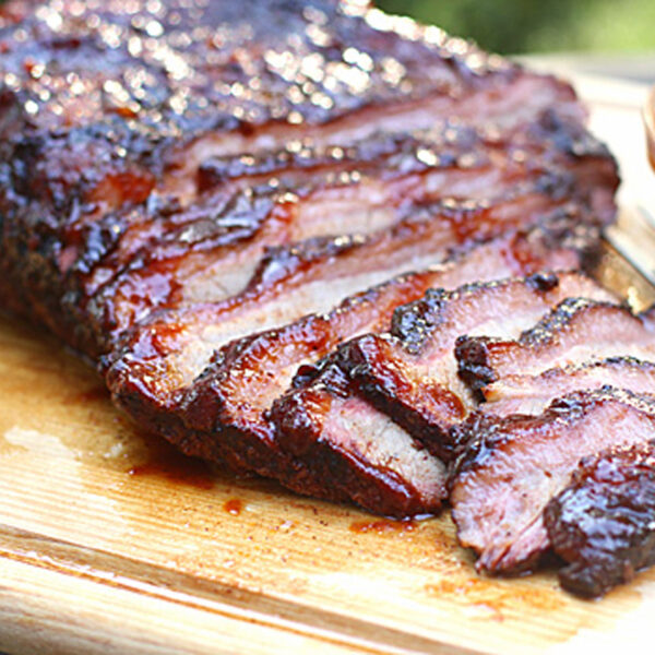 Sliced smoked brisket with a dark, caramelized crust is arranged on a wooden cutting board. A fork and a small bowl of barbecue sauce sit beside the juicy, tender meat.