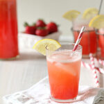 A glass of pink strawberry lemonade with ice, a slice of lemon, and a striped straw sits on a white napkin. In the background are fresh strawberries, a pitcher, and more glasses of the drink.