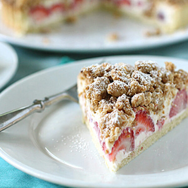 A slice of strawberry crumble cheesecake sits on a white plate with a fork, topped with crumbly streusel and powdered sugar, with the rest of the cake blurred in the background.