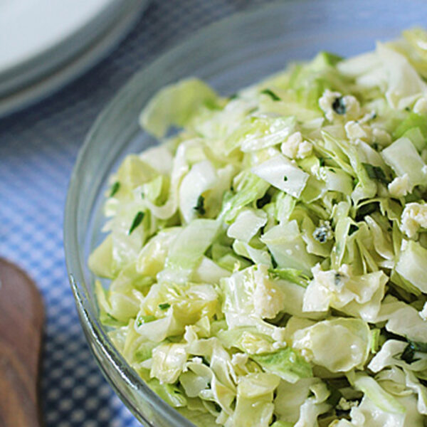 A glass bowl filled with chopped cabbage salad on a blue and white checkered cloth, with wooden salad tongs and stacked white plates in the background.