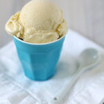 A scoop of vanilla ice cream in a blue cup sits on a folded white cloth napkin, with a white spoon placed beside it.