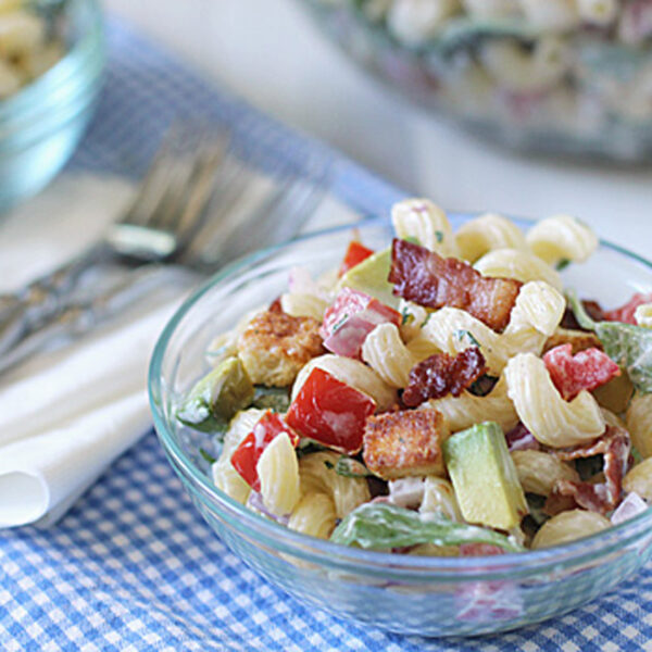 A glass bowl of pasta salad with spiral noodles, bacon, tomatoes, avocado, and lettuce sits on a blue checkered cloth next to a napkin and forks. Another bowl of salad is visible in the background.
