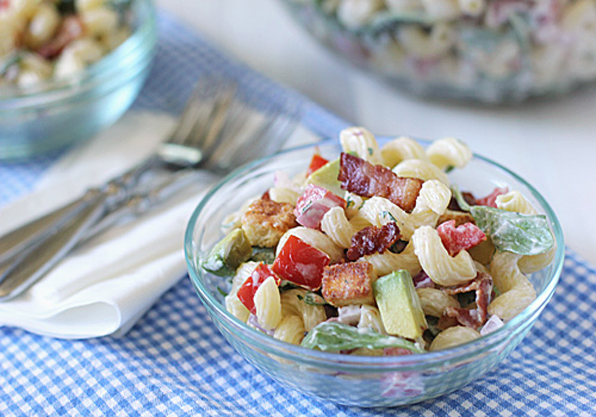 A glass bowl of pasta salad with spiral noodles, bacon, tomatoes, avocado, and lettuce sits on a blue checkered cloth next to a napkin and forks. Another bowl of salad is visible in the background.