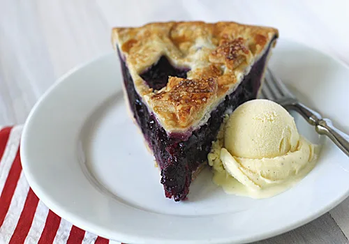 A slice of blueberry pie with golden lattice crust is served on a white plate alongside a scoop of vanilla ice cream, with a fork and a red-striped napkin beside the plate.