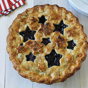 A golden-brown pie with star-shaped cutouts on the top crust, showing dark berry filling. The pie sits on a light wooden surface next to red and white napkins and stacked white plates with silver forks.