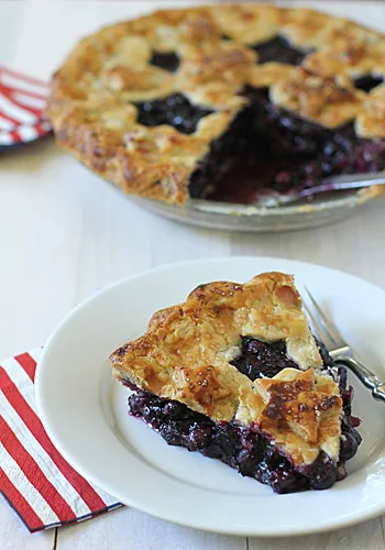 A slice of berry pie with a golden, flaky crust sits on a white plate with a fork. The rest of the pie is in the background. A folded napkin with an American flag design is beside the plate.