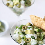 A close-up of ceviche served in a martini glass, garnished with chopped avocado, green olives, cilantro, and two tortilla chips on the side. Another glass with ceviche is blurred in the background.